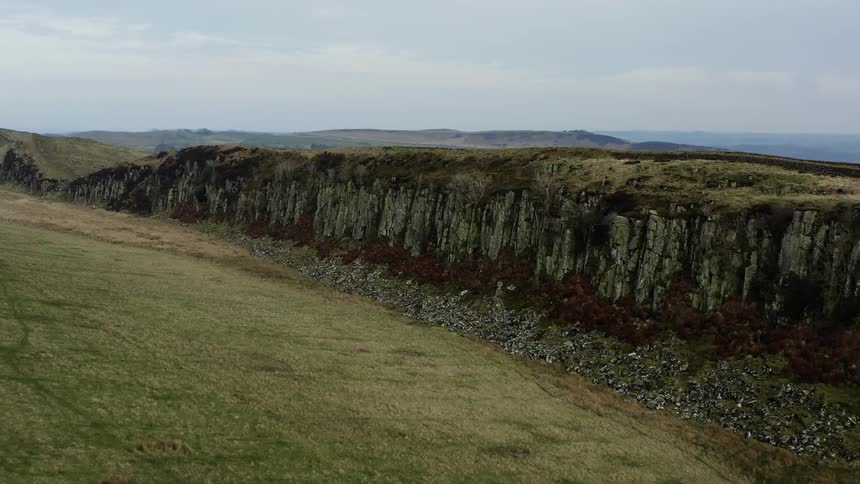 The camera is moving forward over a field and towards a steep rock faced cliff