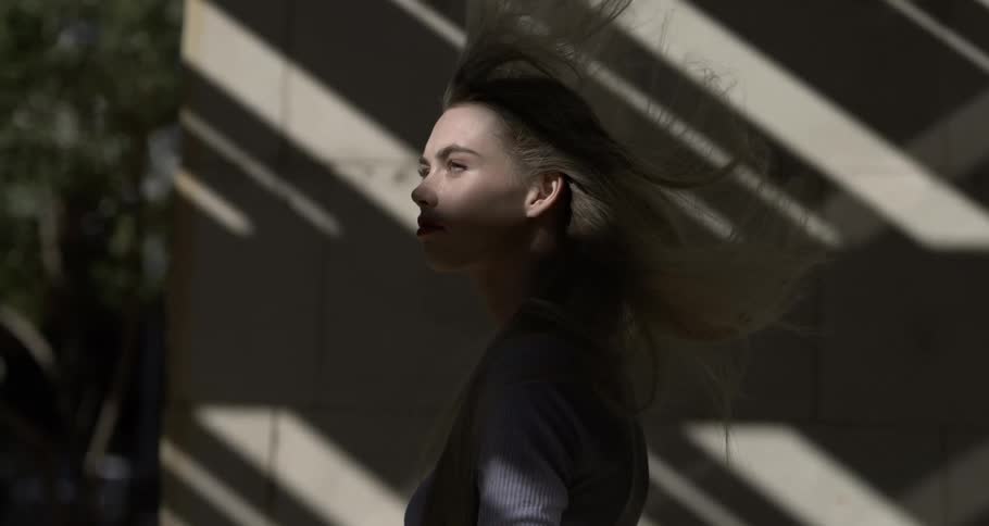 A young woman with long flowing hair walks beneath shadows cast by the buildings awning