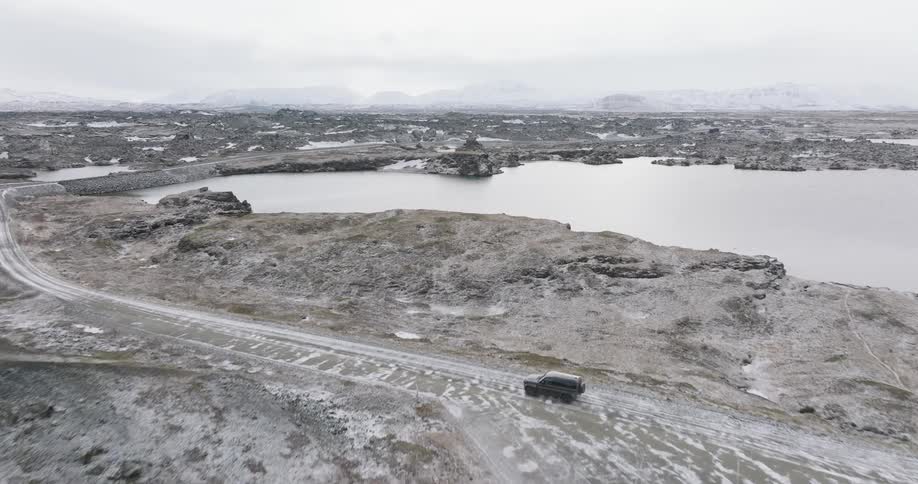 An aerial view of a black car driving down a sandy street and towards the lake there is a large still lake
