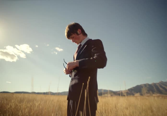 Man in the midst of a field with tall grass wearing a suit carrying a backpack cleaning his glasses and walking