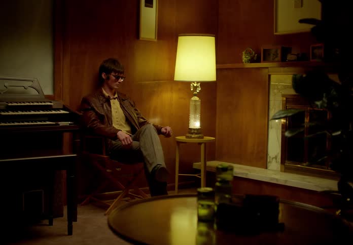 A man sits down on a chair in a living room and puts his sunglasses on