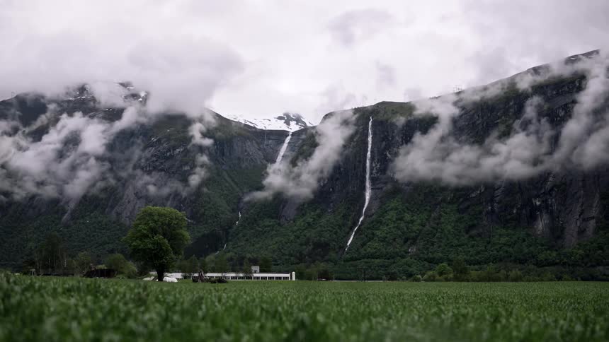 A landscape of a mountain of rocks and some fog clouds and on the ground a lot of green grass