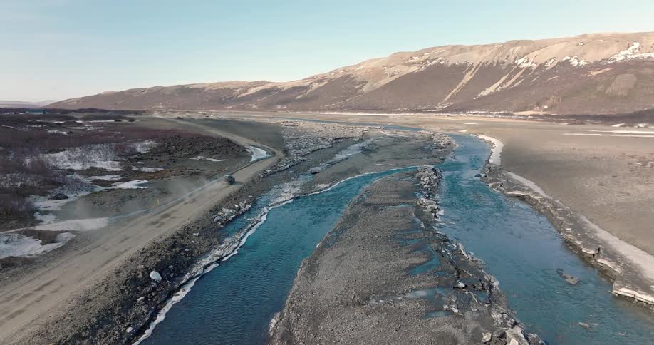 Aerial panorama of a car driving on a dirt road beside a river and mountains