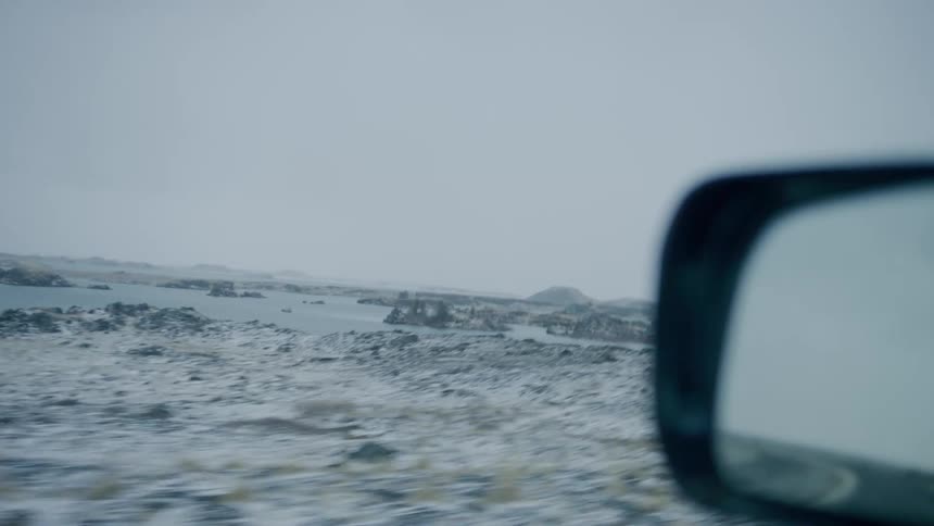 Snow covered terrain beside an icy lake below a pale blue sky from a driving perspective out the drivers side window