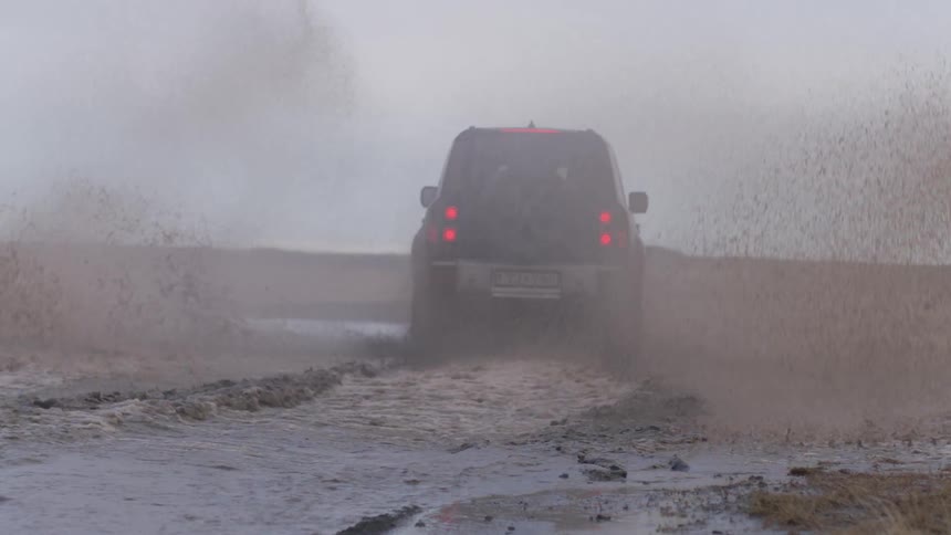A large black car passes over a puddle of muddy water and makes the water rise to the sides