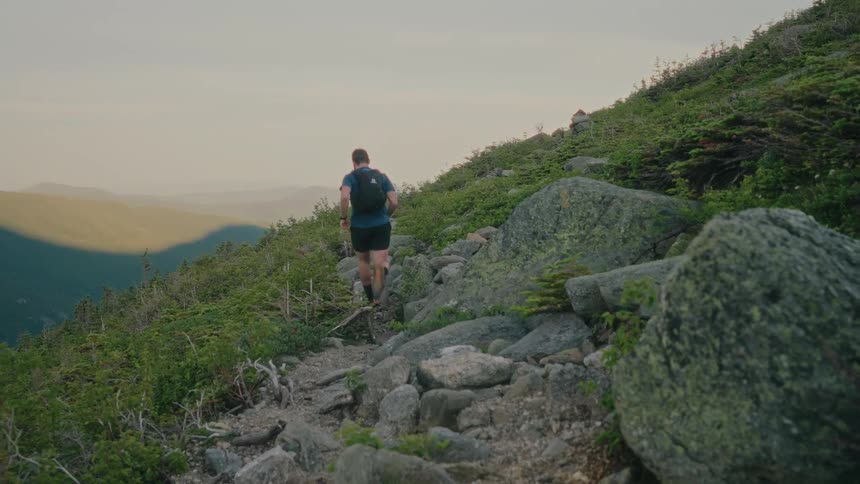 Man running beside large rocks and green grass on the side of a hill with mountains in the distance against a pale sky