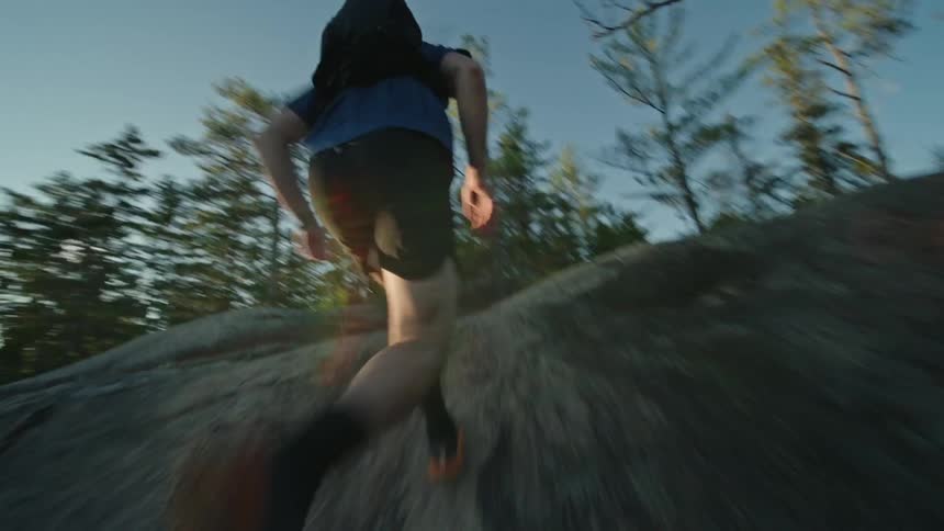 Rear view of a man with a backpack running up a rocky hill surrounded by trees in an outdoor setting