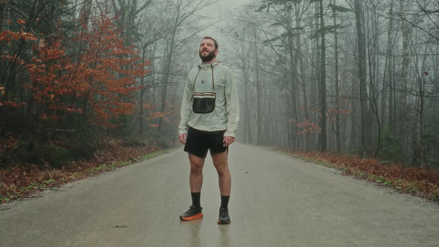 A man stands in the middle of a forest road looking around as it rains