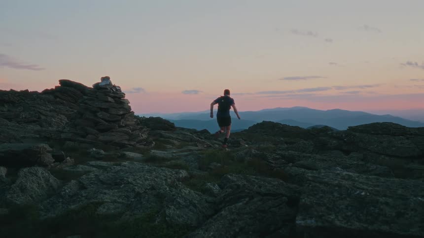 A man with a backpack run along a rocky trail towards a cairn at twilight with a backdrop of a softly lit sky and distant mountains