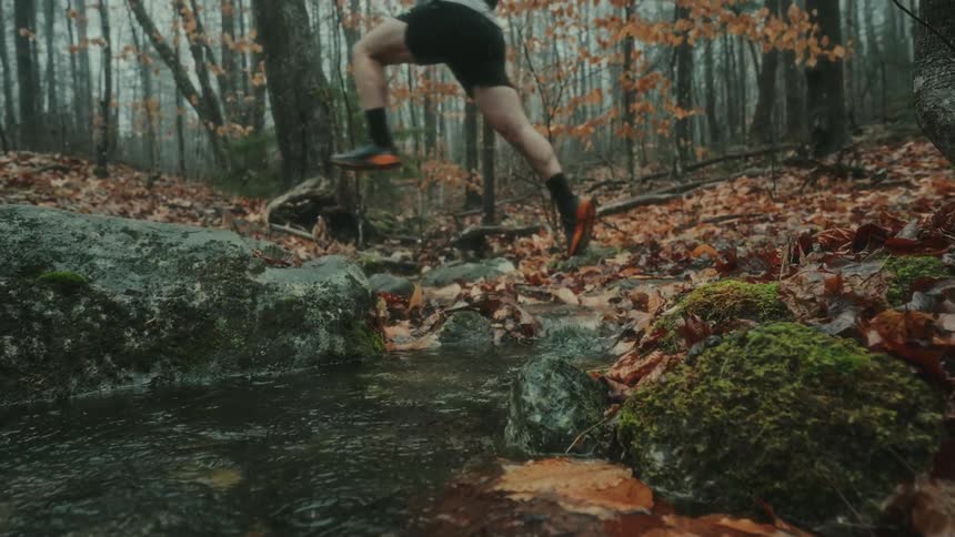A person is jumping over a forest stream surrounded by autumn leaves and mossy rocks