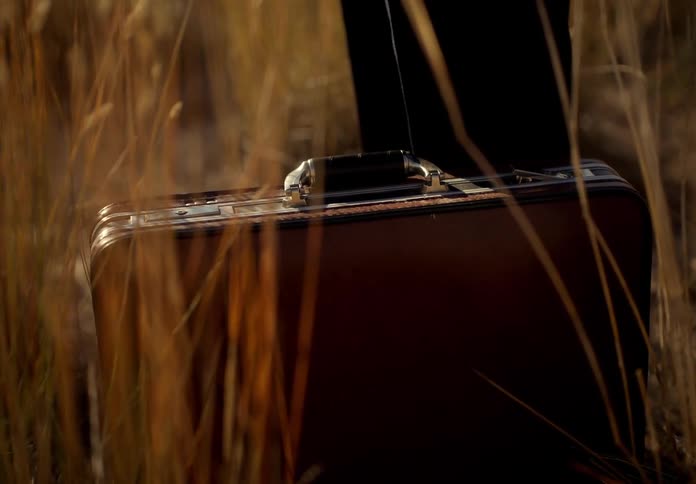 A human hand picking up a briefcase that is on the ground near tall grass