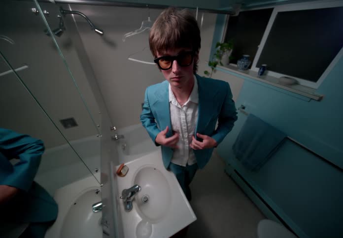 A man in a bathroom puts on sunglasses and preens in the mirror