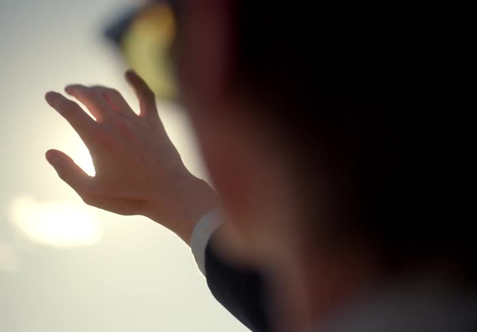 Closeup on the face of a man in sunglasses and a suit raising his hands towards the sun