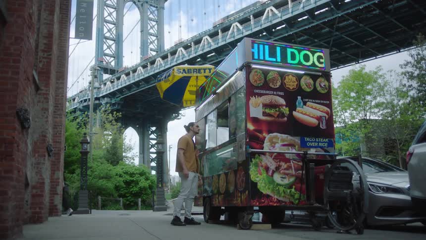 A man is standing looking at a food stall on the street next to a huge bridge