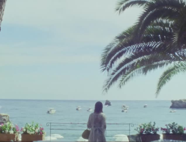 A woman is standing alone watching the boats in the ocean from a walkway on the coast