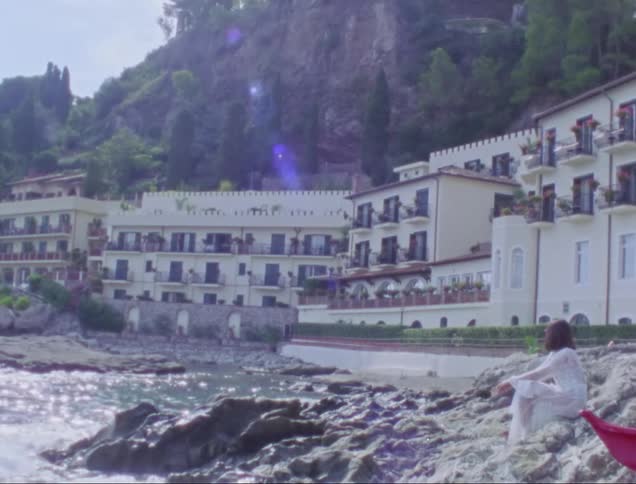 Woman sitting on rocks by the sea on the beach next to a hotel