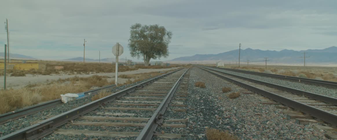 Railway tracks stretch across the land with mountains in the distance under a cloudy sky
