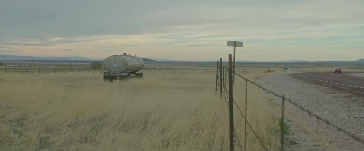 A rural region with dry grass and a stopped truck and next to it a road with a car passing by