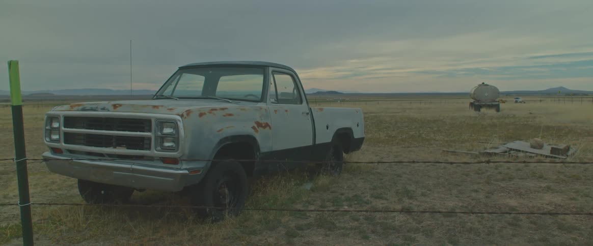 An abandoned pickup truck sits in a large grassy field on a cloudy day