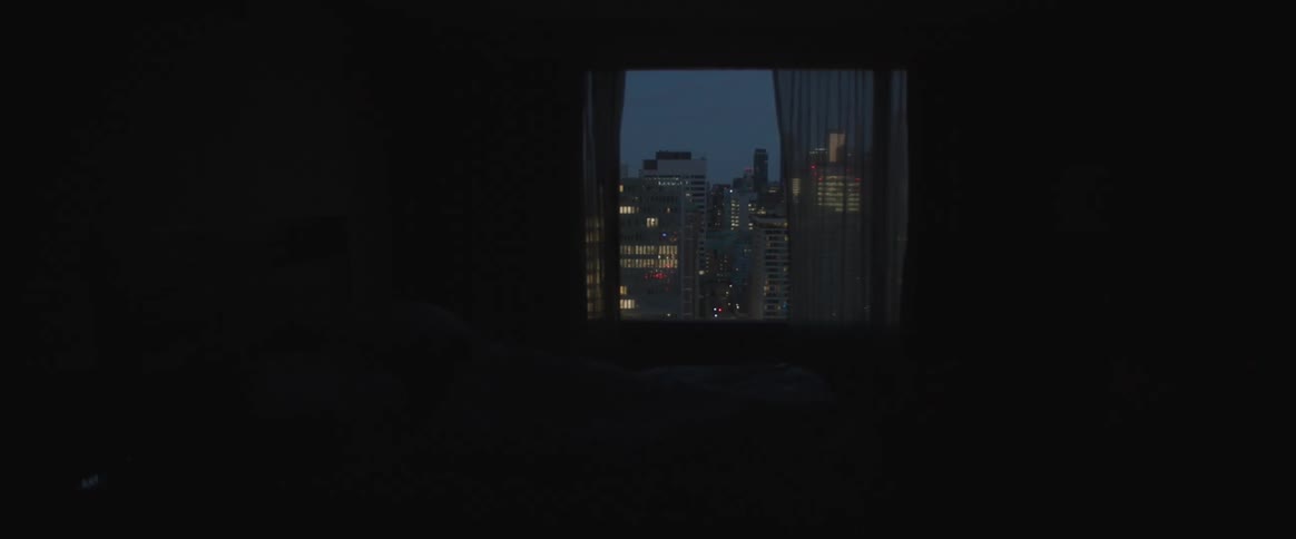 The pitch black room has a window with a view of the city