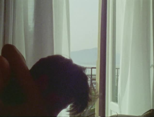 A man and a woman kissing on a bed in front of an open window leading to a balcony