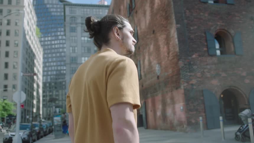 A man with tied hair and a yellow shirt is walking near brick buildings in a city