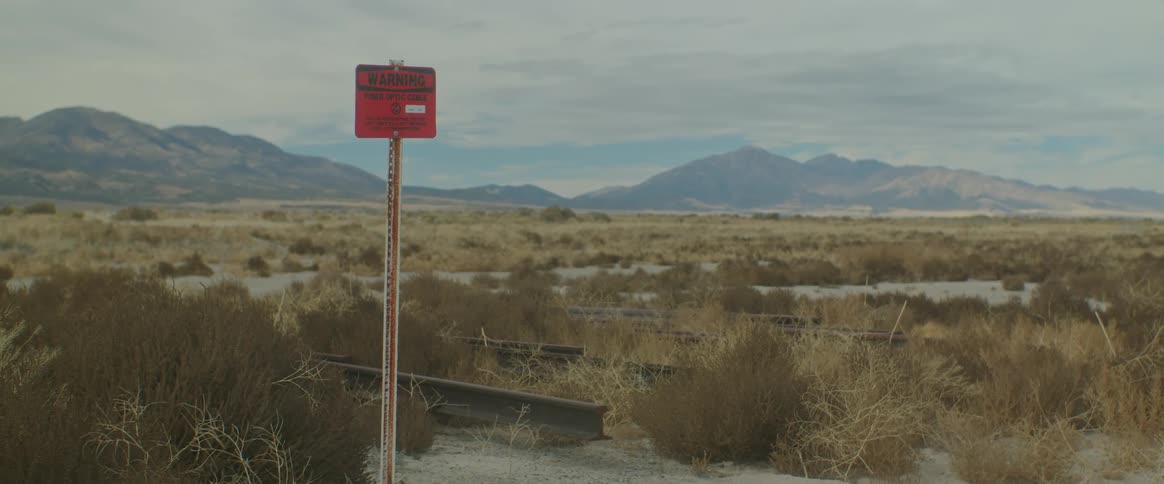 A warning sign on a rusty pole stands in front of a vast expanse filled with tumbleweeds in front of a mountain range