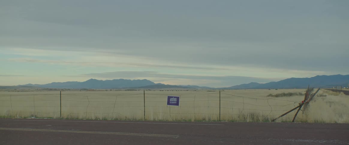 View of a road alongside a fence in a pasture with a chain of mountains on the horizon