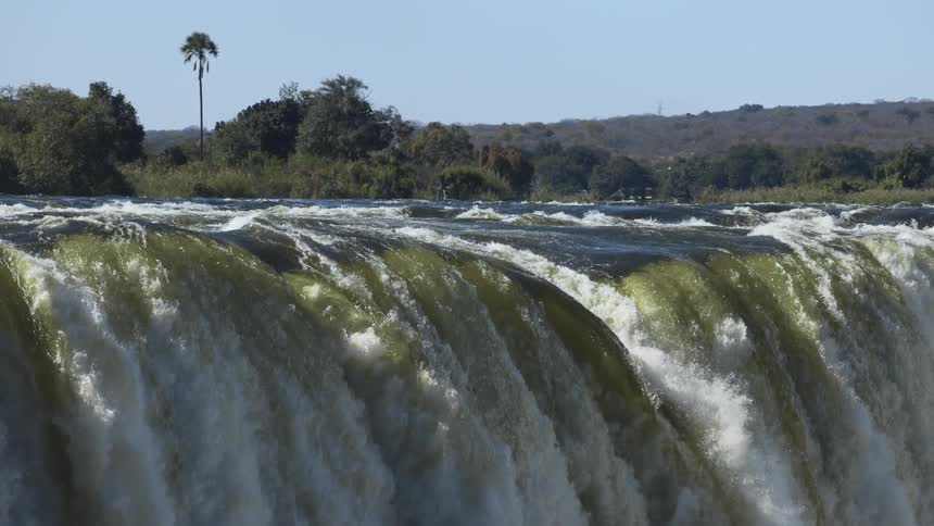 Waterfalls flow over a cliff with trees in the distance