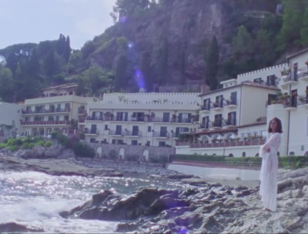 A woman in a white dress is on the sea coast on top of rocks and there are buildings behind her
