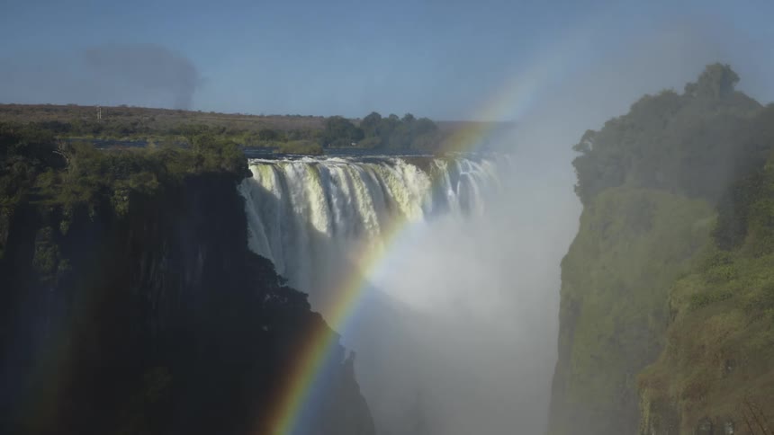 A rainbow over victoria falls in the rainforest of zimbabwe under a bright blue sky