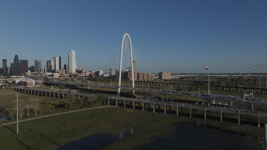 Traffic crosses suspension bridge over a river passing a texas flag on route to and from city downtown