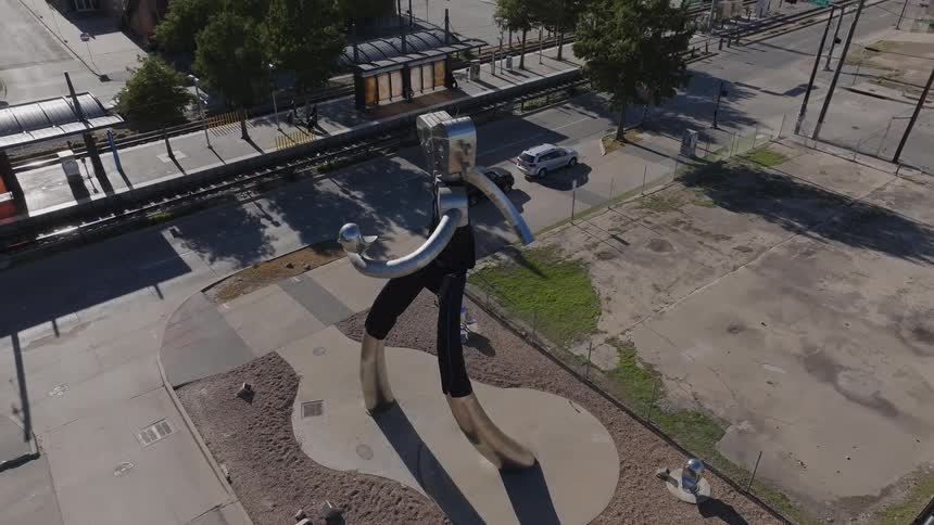 Tall modern metal art sculpture of a slinky arm and legged robot at the edge of the city with the skyline and blue sky behind it on a sunny day