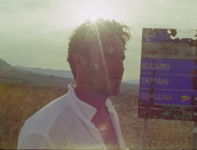 Man stands next to damaged road sign in field on bright windy day