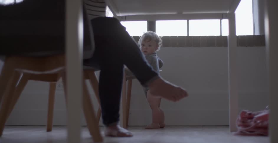 A baby is pushing a chair by a window