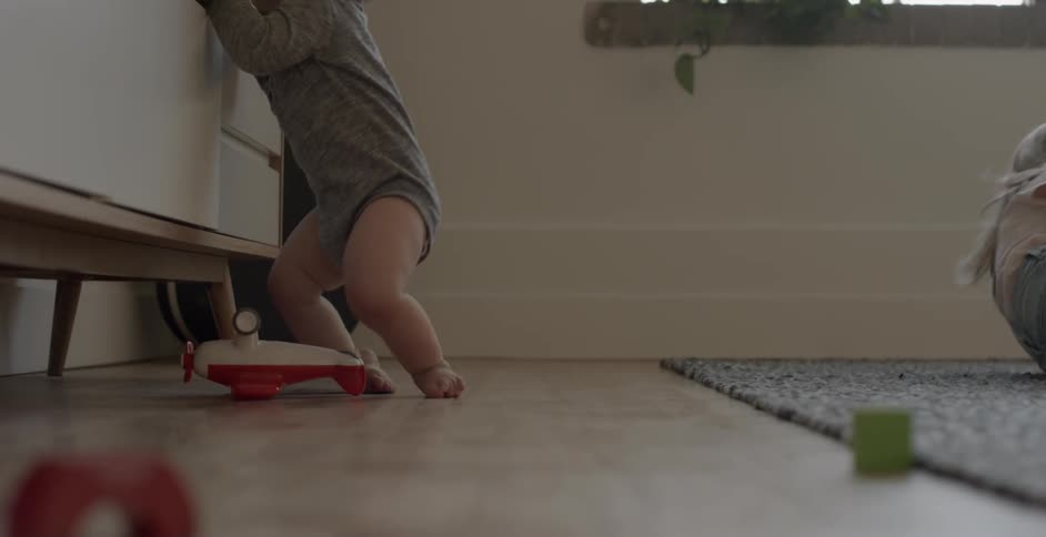 A baby is crawling on dresser as another child lays on the carpet