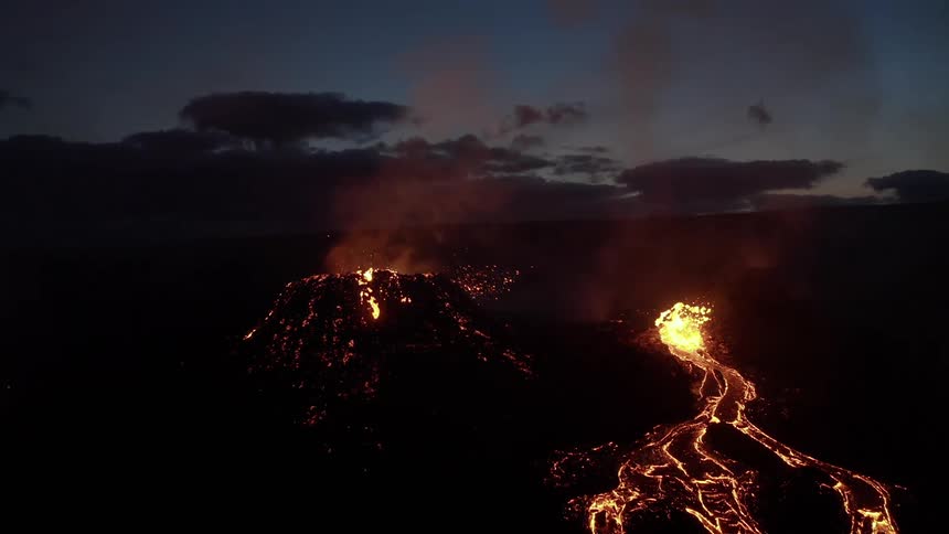 Molten hot lava glows in the darkness as it splashes from a volcano and smoke rises as lava flows