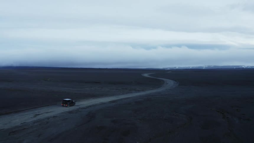 A jeep with taillights on driving on a winding dirt highway across flat land under a wide open cloudy sky at nightfall