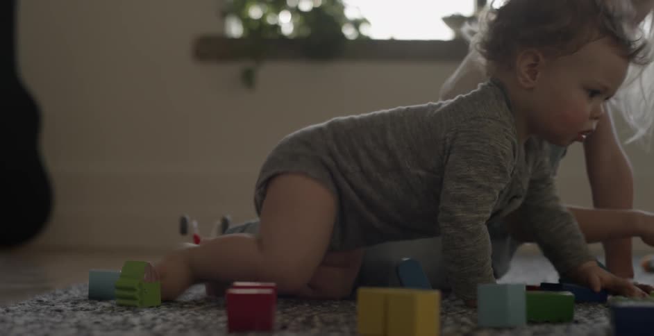 A toddler crawls on a rug while a child plays with toys