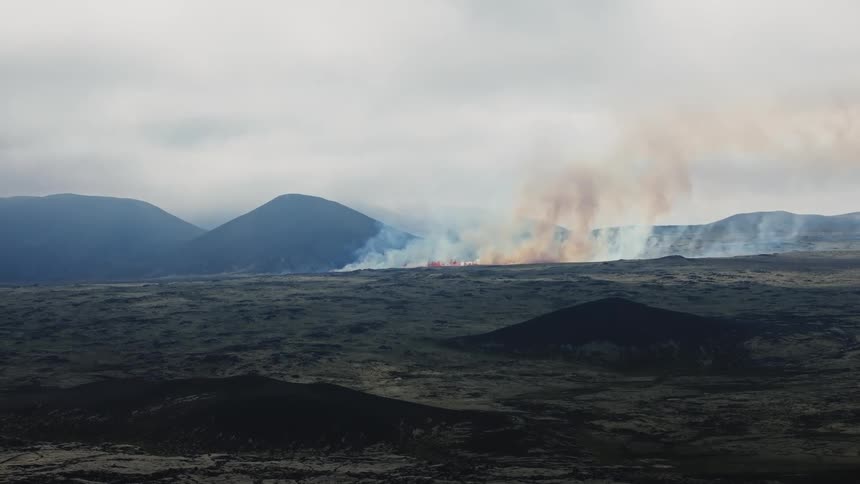 Landscape of volcano eruption with little lava and lots of smoke next to a mountain range during day time