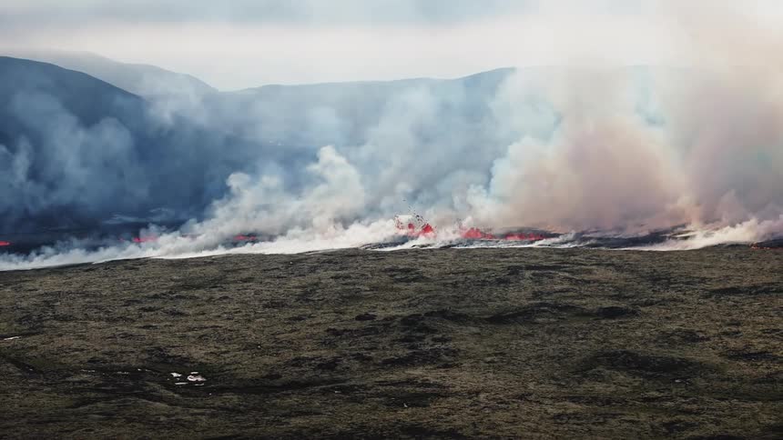 Aerial of active volcanic fissure expelling lava and plumes of white smoke with mountains outlining the horizon