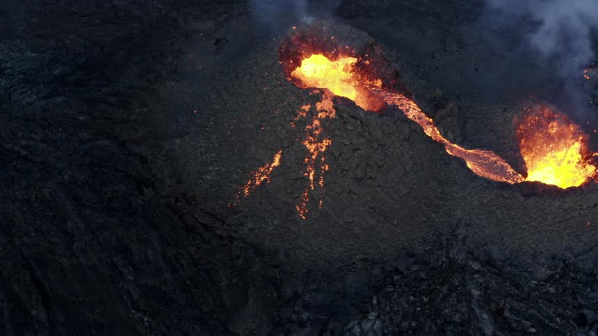 Molten hot lava glows in the darkness as it splashes from an erupting volcano and smoke rises as lava flows