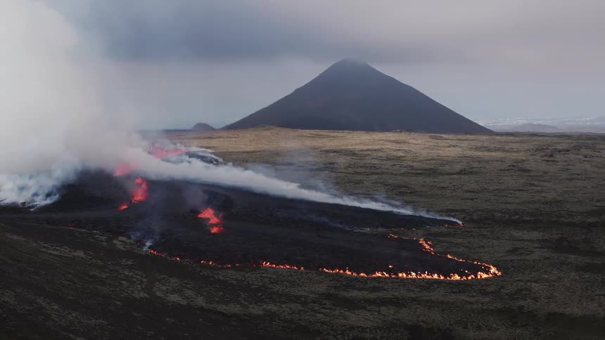 Fire around a black lava flow with rolling smoke on a sloping terrain with a mountain and cloudy sky in the background