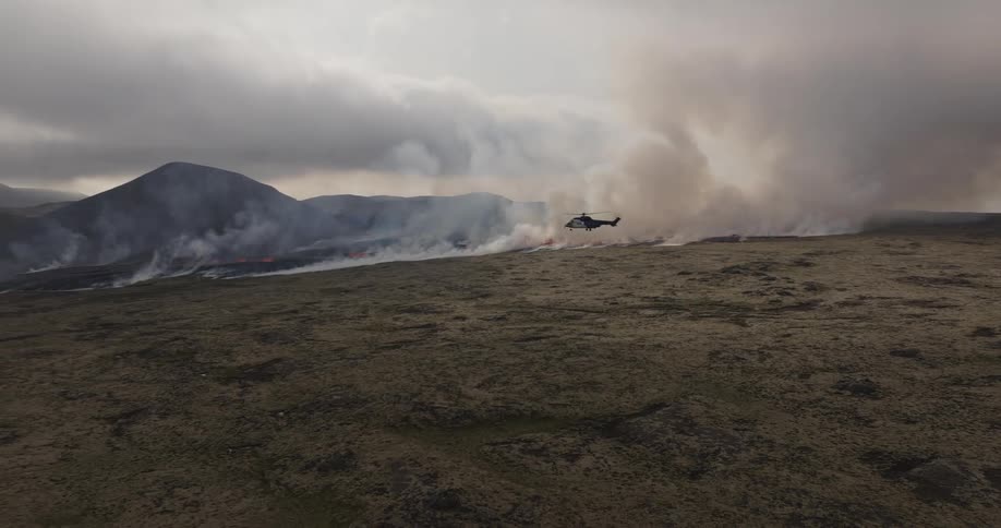 Helicopter flies close to wildfire burning near base of hills on an overcast day