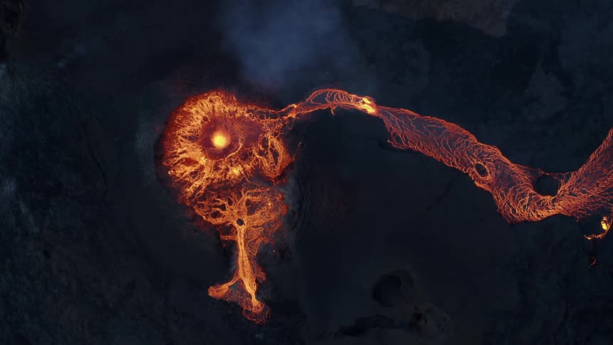 Glowing molten hot lava splashes from an erupting volcano and flows down the sides and across the land in the darkness
