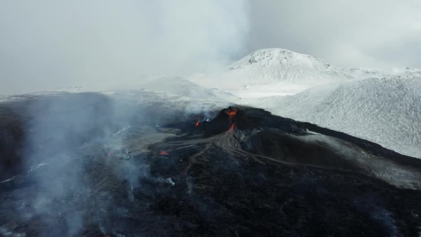 Aerial landscape of volcano erupting next to a mountain range covered in snow during day time