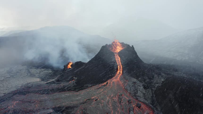 Molten hot lava glows as it splashes and flows from an erupting volcano and snow flies among smoke in the air