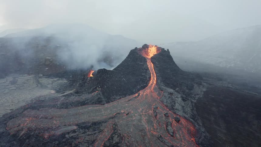 Molten hot lava glows as it splashes and flows from an erupting volcano and snow flies in the air
