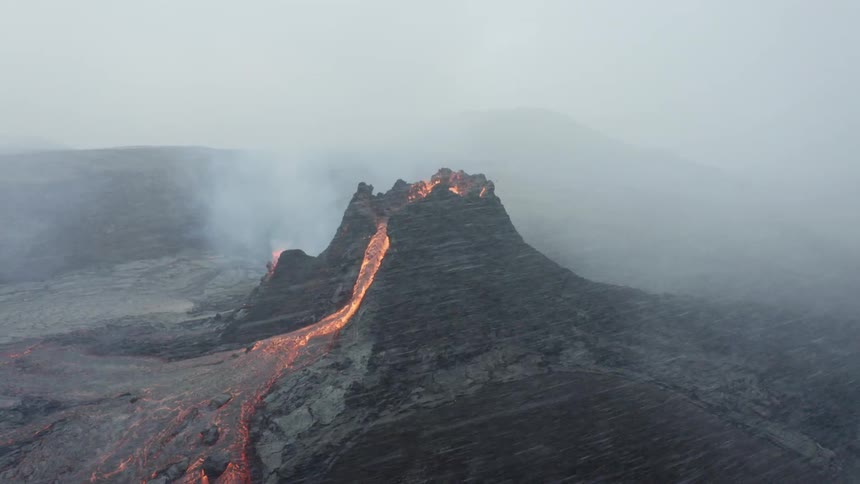 Molten hot lava glows as it splashes from an erupting volcano and flows across the land and snow flies among smoke in the air