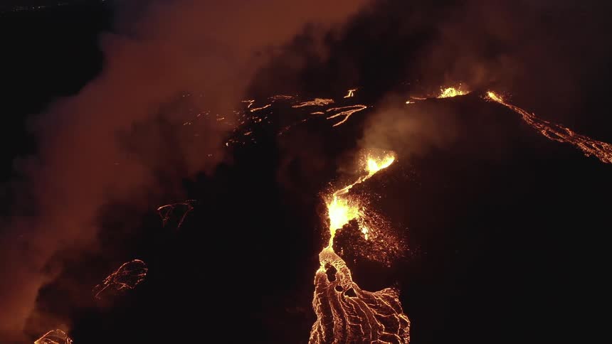 Molten hot lava glows in the darkness as it splashes from an erupting volcano and smoke rises as lava flows